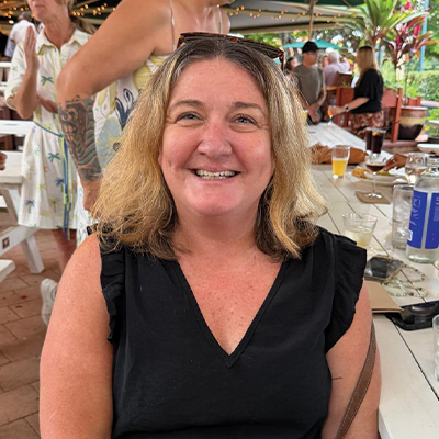 A woman in a black sleeveless top sits at a white wooden table in an outdoor café, smiling warmly at the camera. The setting is lively, with string lights, tropical plants, and other diners visible in the background. Drinks and food are spread across the table, capturing a relaxed, social atmosphere in a warm climate.
