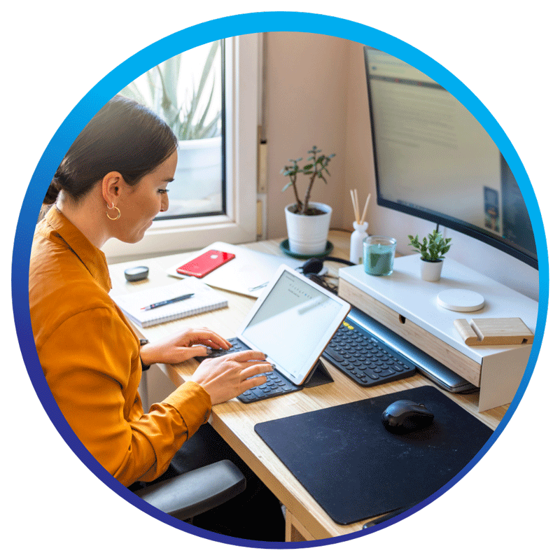 "A person is working at a desk with a laptop and a large monitor. The desk is organized with various items including a keyboard, mouse, notepad, pen, smartphone, and several potted plants. The person is typing on the laptop, which is placed on a stand, and the monitor displays some content. The workspace is well-lit with natural light coming from a window.
