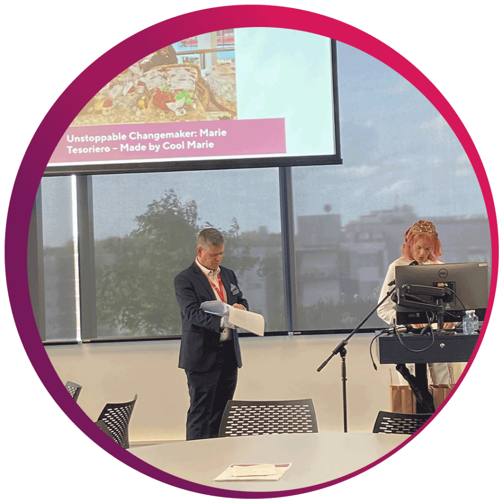 Two individuals are in a professional setting, likely during a presentation or conference. One person is standing and holding a piece of paper, while the other stands behind a podium with a computer and microphone. The background features a large window showcasing trees and buildings. A presentation slide is visible on the screen, reading: 'Unstoppable Changemaker: Marie Tesoriero – Made by Cool Marie.