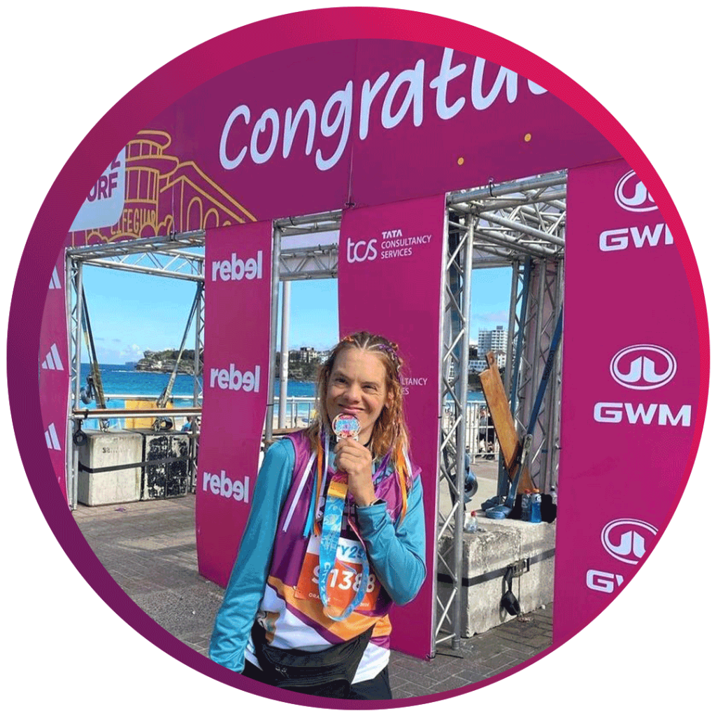 A person is standing in front of a pink backdrop with the word 'Congratulations' partially visible at the top. The backdrop features logos for 'TCS Tata Consultancy Services,' 'rebel,' and 'GWM.' The person is wearing a blue and purple athletic outfit with a race bib number '21338' and is holding a medal. The background shows a coastal scene with water and buildings.