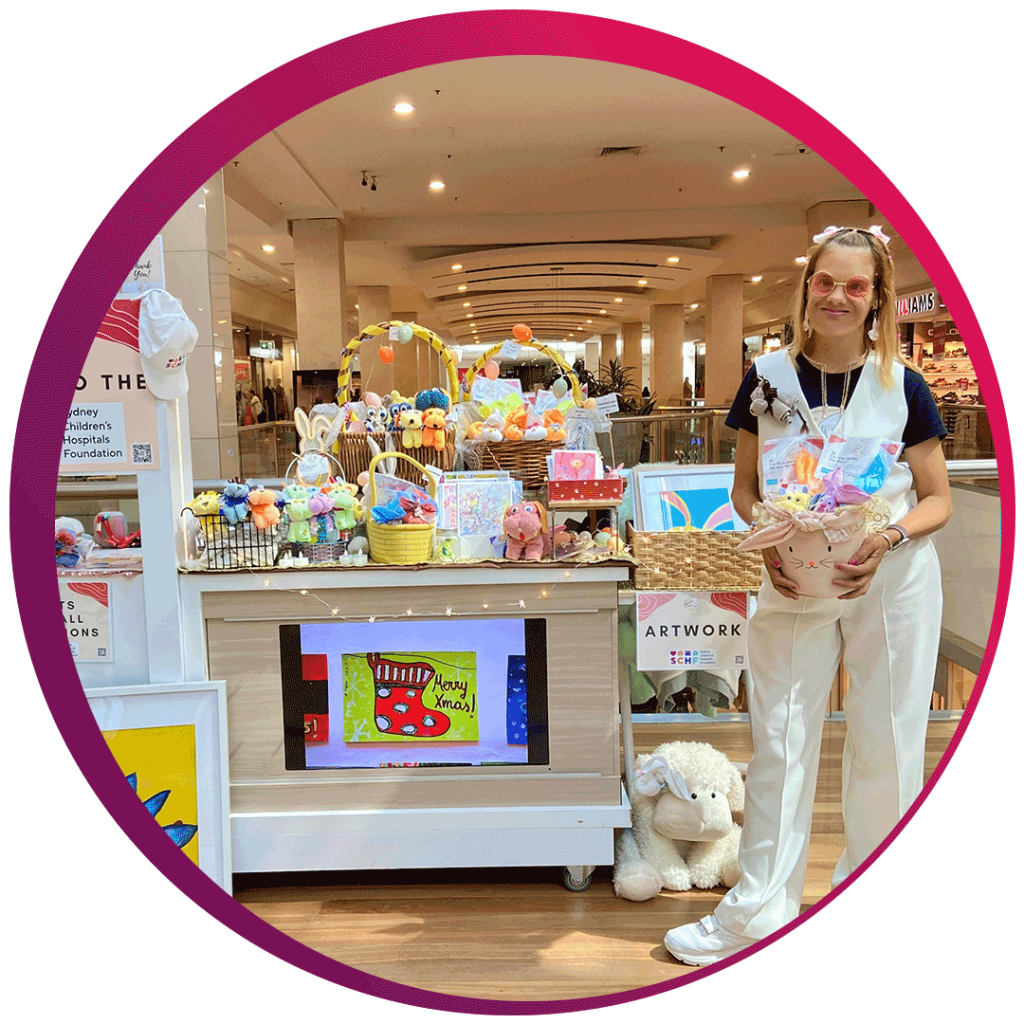 A person is standing next to a display booth in a shopping mall. The booth is filled with various colorful stuffed animals, toys, and artwork. The person is holding a basket filled with toys and is wearing a white outfit. There is a sign on the booth that reads 'Merry Xmas!' with a drawing of a Christmas stocking. The booth also has a sign that says 'ARTWORK' and mentions the Sydney Children’s Hospitals Foundation. The background shows a busy shopping mall with people walking and various stores.