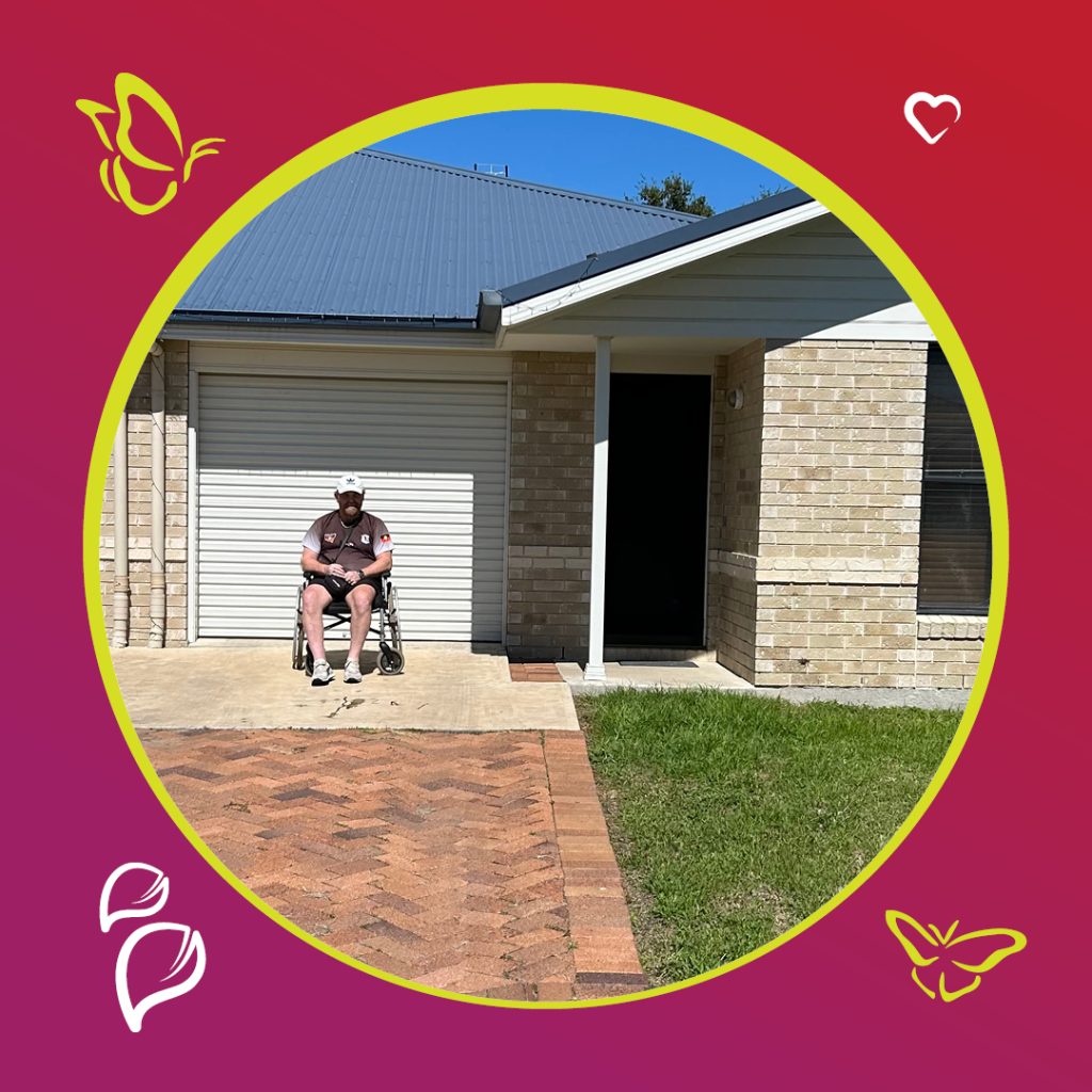 A person sits in a wheelchair on a concrete driveway in front of a house with beige brick walls and a blue metal roof. The house features a closed garage door and a covered entrance. The driveway is bordered by a brick pathway and a grassy area. The image has a circular frame with a gradient background transitioning from red to purple, decorated with illustrations of butterflies, leaves, and a heart." Does this align with what you need? I can refine it further if you'd like a more tailored description!