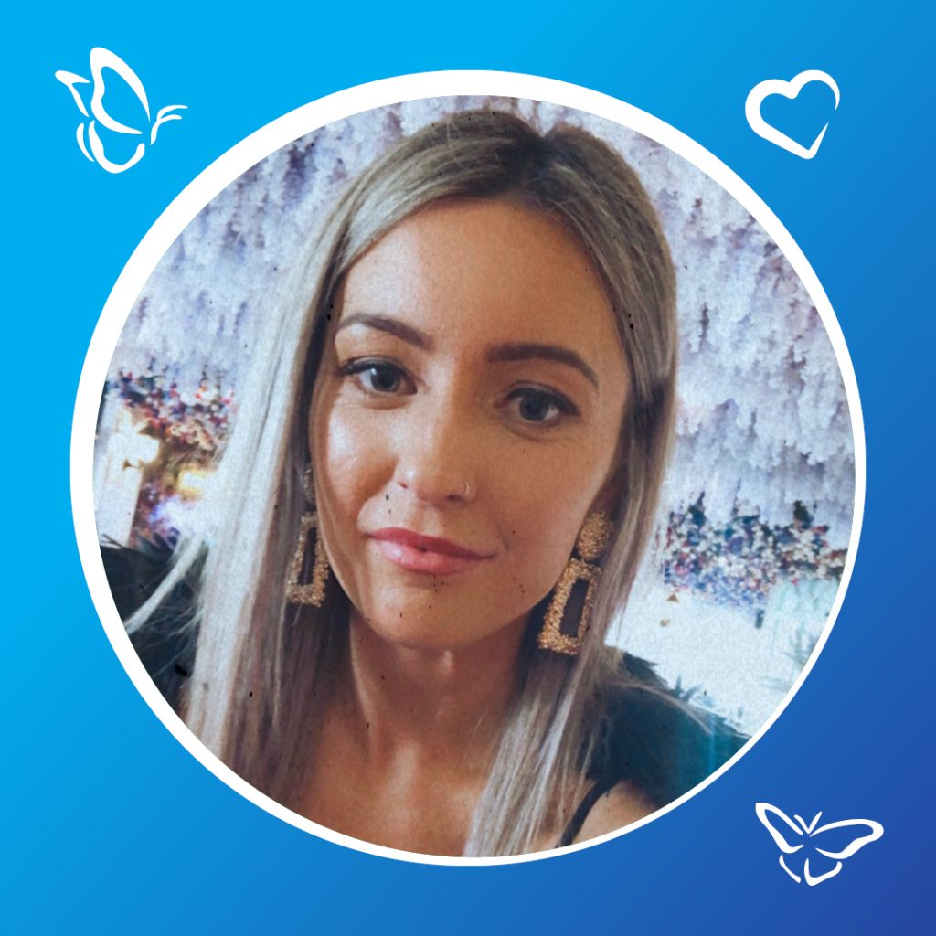 A person with long, straight, light-colored hair wears a black top, standing in front of a floral arrangement with purple and white flowers. The image is framed by a blue circular border, decorated with white butterfly and heart icons in the corners." Let me know if you'd like any adjustments to match your preferred style or accessibility needs!