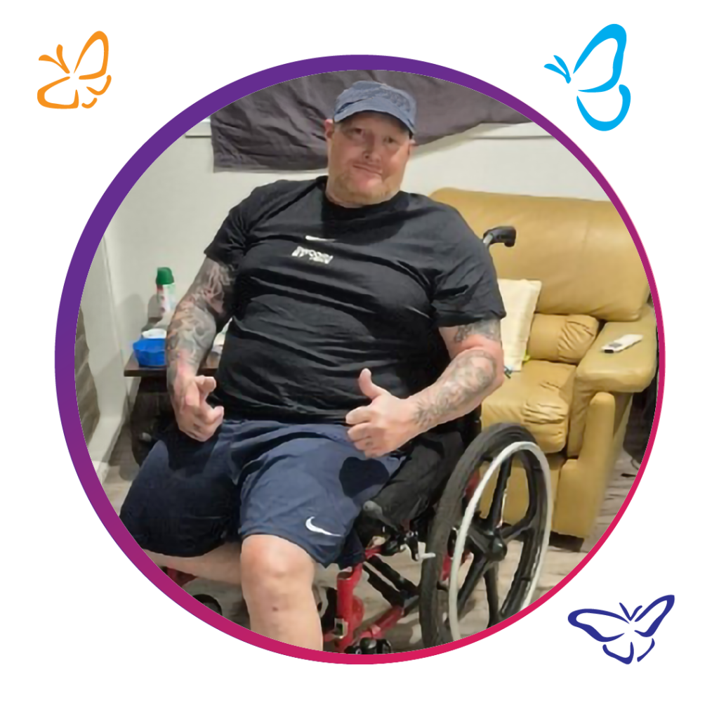 A person in a wheelchair smiles and gives a confident thumbs-up with both tattooed arms. They wear a black t-shirt and blue shorts. Behind them, a cozy yellow armchair holds a pillow and a remote, while a small table nearby displays a green bottle among other items. The image is framed by a purple circle, adorned with lively orange and blue butterfly illustrations, adding a sense of vibrancy.