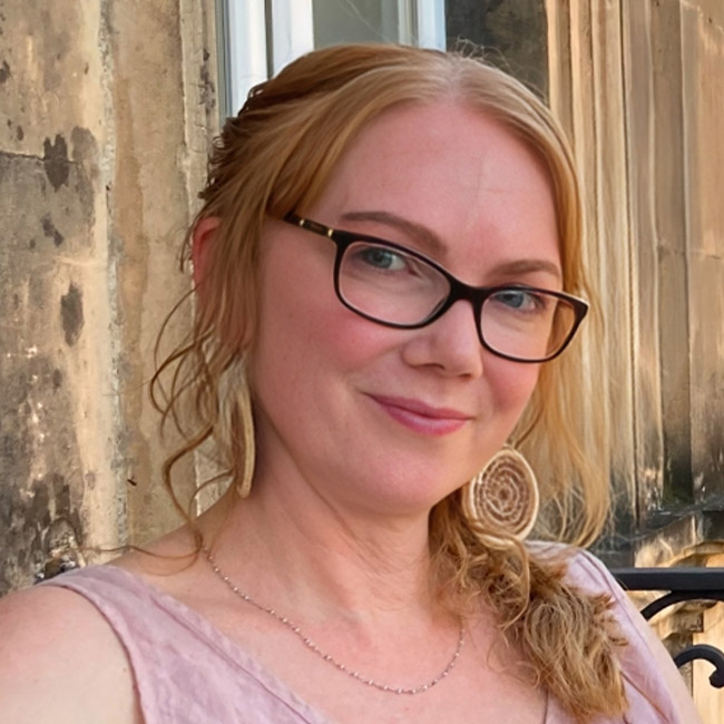 A picture of a smiling women with long blond hair, black framed glasses in a pink top smiling at the camera.
