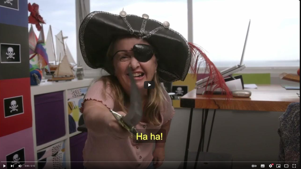 A person dressed as a pirate, wearing a decorated hat with a red feather, confidently points a curved sword toward the camera. Their stance is playful yet commanding. The backdrop features colourful storage boxes, a desk with a laptop and miscellaneous items, and a wall adorned with black squares displaying white skull and crossbones symbols. The text 'Ha ha!' appears at the bottom, emphasising the character’s exuberant energy.