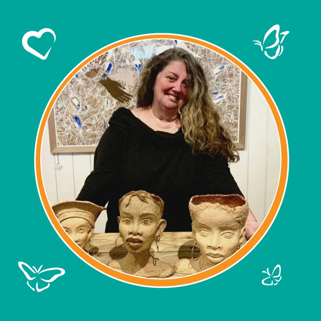 An image of a woman standing behind a table that has 3 human head busts. The woman is smiling and behind her is a detailed painting. The image is inside a circle with an orange boarder, sitting on a green background. In the corners of the image are icons of 3 butterflies and 1 heart.