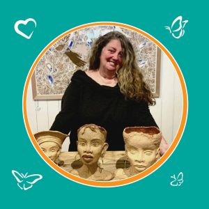 An image of a woman standing behind a table that has 3 human head busts. The woman is smiling and behind her is a detailed painting. The image is inside a circle with an orange boarder, sitting on a green background. In the corners of the image are icons of 3 butterflies and 1 heart.
