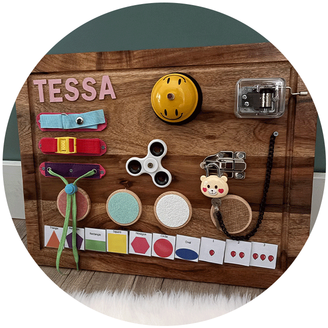 A wooden board with various different items attached to it. sitting on a wooden floor in front of a white fuzzy rug against green wall with white skirting board. The items on the board include; a bell, buckles, texture pads, a latch with a teddy bear head shaped pad lock, cards with different colours and shapes on them and a music box.