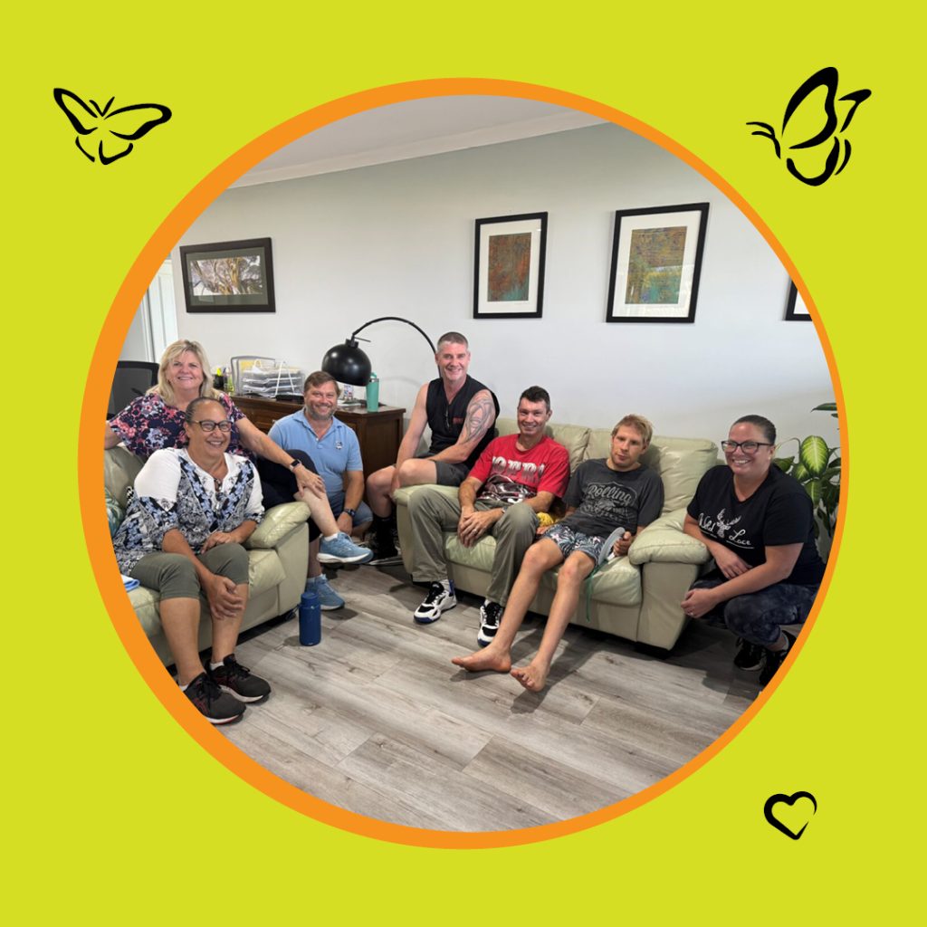A picture of a group of people sitting in a lounge room on lounge chairs smiling at the camera. The image is in a circle frame with an orange boarder sitting on a yellow-green background with icons of butterflies and a heart in three of the corners.