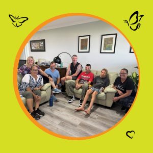 A picture of a group of people sitting in a lounge room on lounge chairs smiling at the camera. The image is in a circle frame with an orange boarder sitting on a yellow-green background with icons of butterflies and a heart in three of the corners.