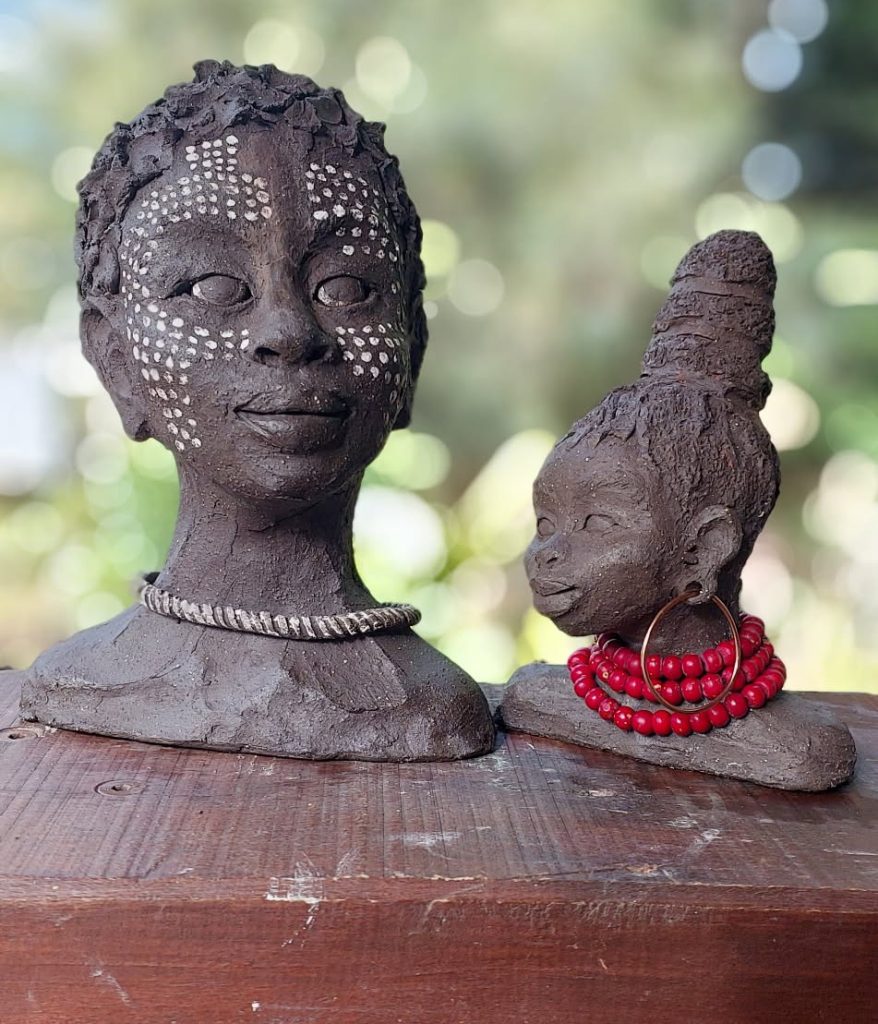 A image of 2 sculptures of female human heads, both are styled in the manner of African indigenous cutlers the larger one has white dots painted on her face with a simple braded necklace and short hair. the other smaller on has large hoop earrings with 3 bright red beaded necklaces. Her hair is a large top bun that looks to be bound with rope. Both sculptures are un-varnished and have a natureal earth tone to them. they are sitting on a wooden table with the background being out of focus greenery.