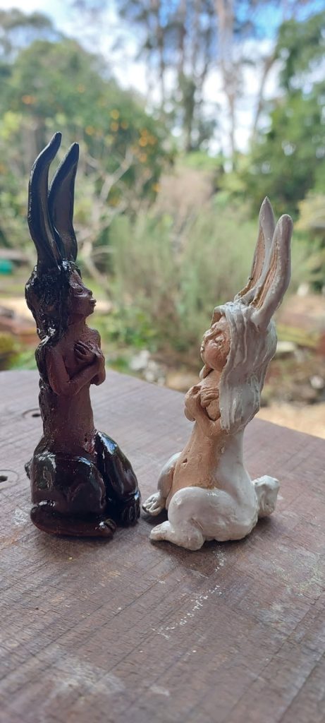 An image of 2 statues 1 black and 1 white, both are of rabbit like creatures sitting upright, however that have human faces and upper bodies with their human arms clasped over their hearts. The statues are sitting on a wooden table with a nature background of shrubbery and trees
