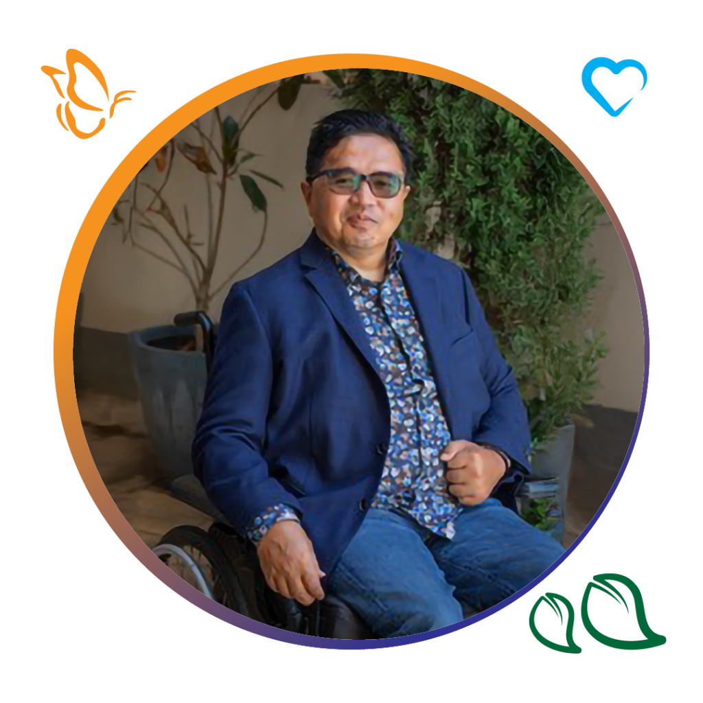 An image for a man smiling art the camera, he is wearing a navy suit jacket and colourful patterned button up shirt and a pair of blue jeans. He has glasses and is sitting in a wheel chair. in the background you can see a concert floor and wall as well as two potted plants. The image is inside a circle with an orange and dark blue boarder. with icons of a butterfly, heart and leaves around the outside of the circle.