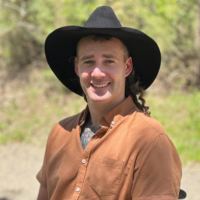 A picture of a man smiling to camera wearing an orange shirt and a wide brim black hat. outside in a forest area.