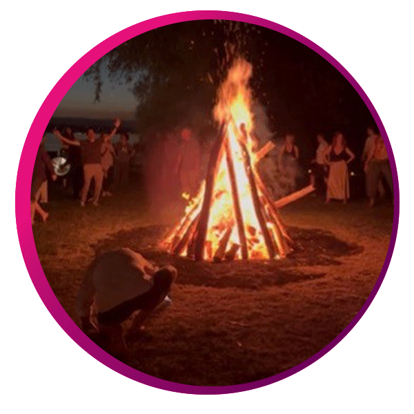 An image of a bonfire with at night time with people standing around the image smiling.