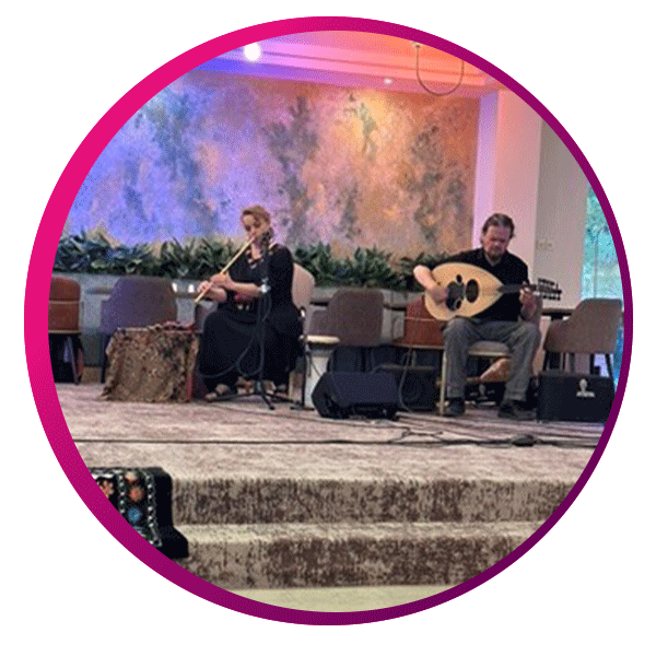 Indoor musical performance featuring two seated musicians on a softly lit stage. One plays a bowed string instrument, possibly a kamancheh, while the other plays an oud, evoking traditional Middle Eastern melodies. Behind them, a textured wall with abstract art and a row of green plants adds warmth and depth. Several empty chairs suggest a larger ensemble or upcoming performers. The setting feels intimate yet formal, with carpeted steps leading to the stage, all framed within a circular pink gradient border that enhances the contemplative mood.