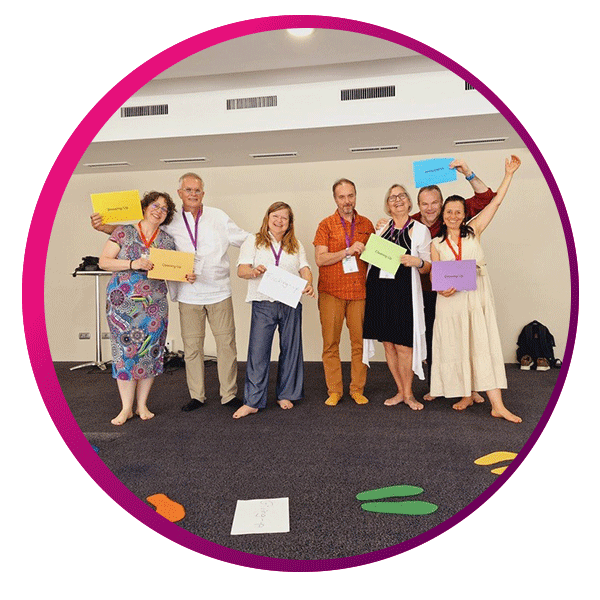 Group of eight adults standing in a semi-circle on a carpeted floor, smiling and holding colorful paper signs with text. Dressed in casual to semi-formal attire, some are barefoot, suggesting comfort and informality. The background includes a white wall with air vents and a table with miscellaneous items. On the floor, vibrant cutouts shaped like footprints and other forms hint at a playful or interactive group activity. The image is framed in a circular border with a soft pink gradient, enhancing the sense of connection and shared experience.