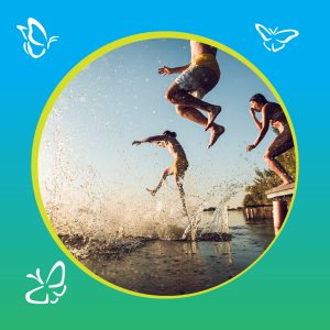 Three people leap joyfully off a wooden dock into a sunlit lake, their bodies mid-air and water splashing dramatically around them. The golden light catches suspended droplets, adding a sense of motion and warmth. Behind them, trees line the calm water, evoking a peaceful summer setting. The image is framed in a circular border with a blue-to-green gradient, accented by delicate white butterfly illustrations in three corners—suggesting whimsy and freedom.
