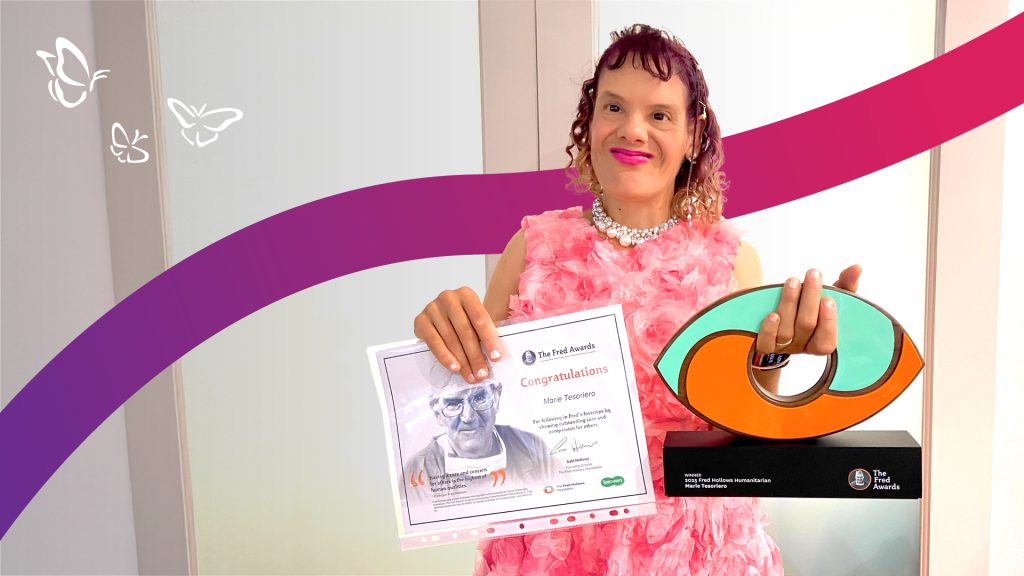 A smiling person in a pink floral dress and pearl necklace holds a certificate in one hand and a vibrant, abstract trophy in the other. The certificate congratulates Marie Tesoriero for winning The Fred Awards for Outstanding Achievement in Reducing the Harms of Drugs, Alcohol and Gambling. The trophy base reads “2023 Fred Hollows Humanitarian.” Behind them, a backdrop of purple and red curved lines with white butterflies adds a celebratory and uplifting tone.