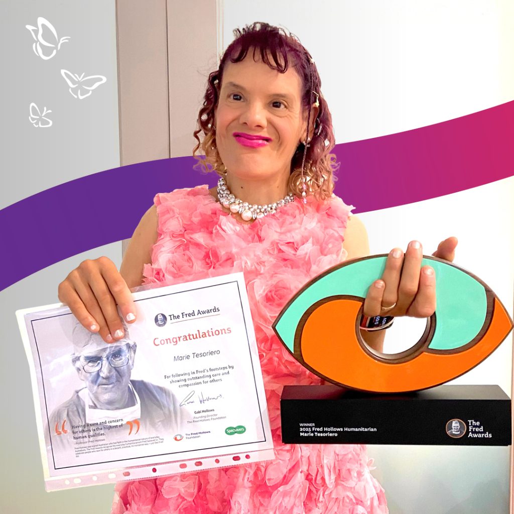 A smiling person in a pink floral dress and pearl necklace holds a certificate in one hand and a vibrant, abstract trophy in the other. The certificate congratulates Marie Tesoriero for winning The Fred Awards for Outstanding Achievement in Reducing the Harms of Drugs, Alcohol and Gambling. The trophy base reads “2023 Fred Hollows Humanitarian.” Behind them, a backdrop of purple and red curved lines with white butterflies adds a celebratory and uplifting tone.