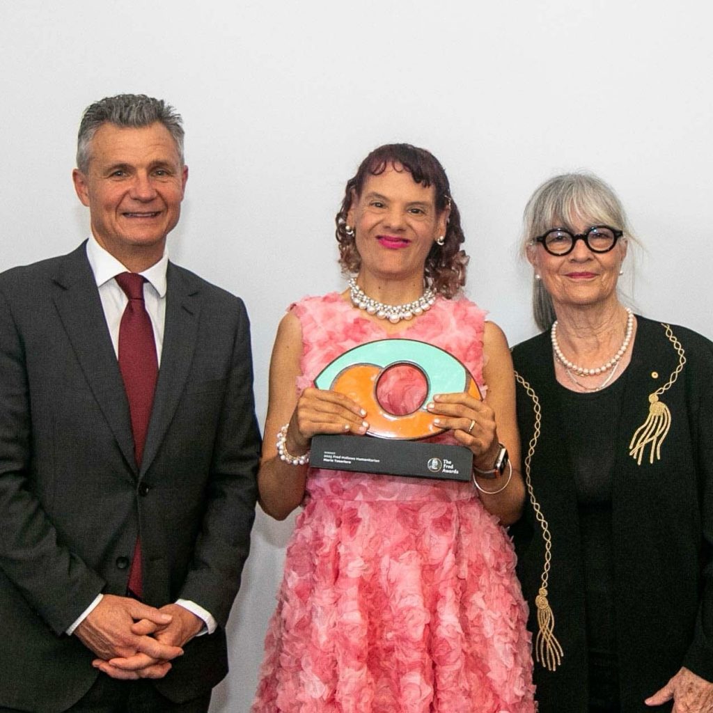 Three people stand side by side against a plain white background. The person in the center, wearing a pink floral dress and pearl jewelry, holds a colorful circular award with orange and green segments. To the left, a person in a dark suit with a red tie smiles warmly. On the right, a person in a black outfit with gold accessories and large round glasses adds a stylish contrast. The scene conveys a moment of recognition and celebration.