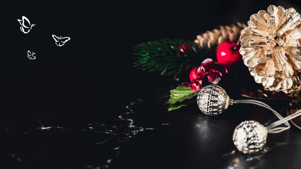 A festive holiday arrangement displayed on a black marble surface, featuring a large gold-painted pinecone and a natural brown pinecone surrounded by artificial red berries, green pine needles, and textured silver baubles on clear wires. Three white butterfly illustrations of varying sizes float on the left side, adding a delicate, whimsical touch. The dark background enhances the brightness and reflective quality of the decorations, creating a striking seasonal contrast.