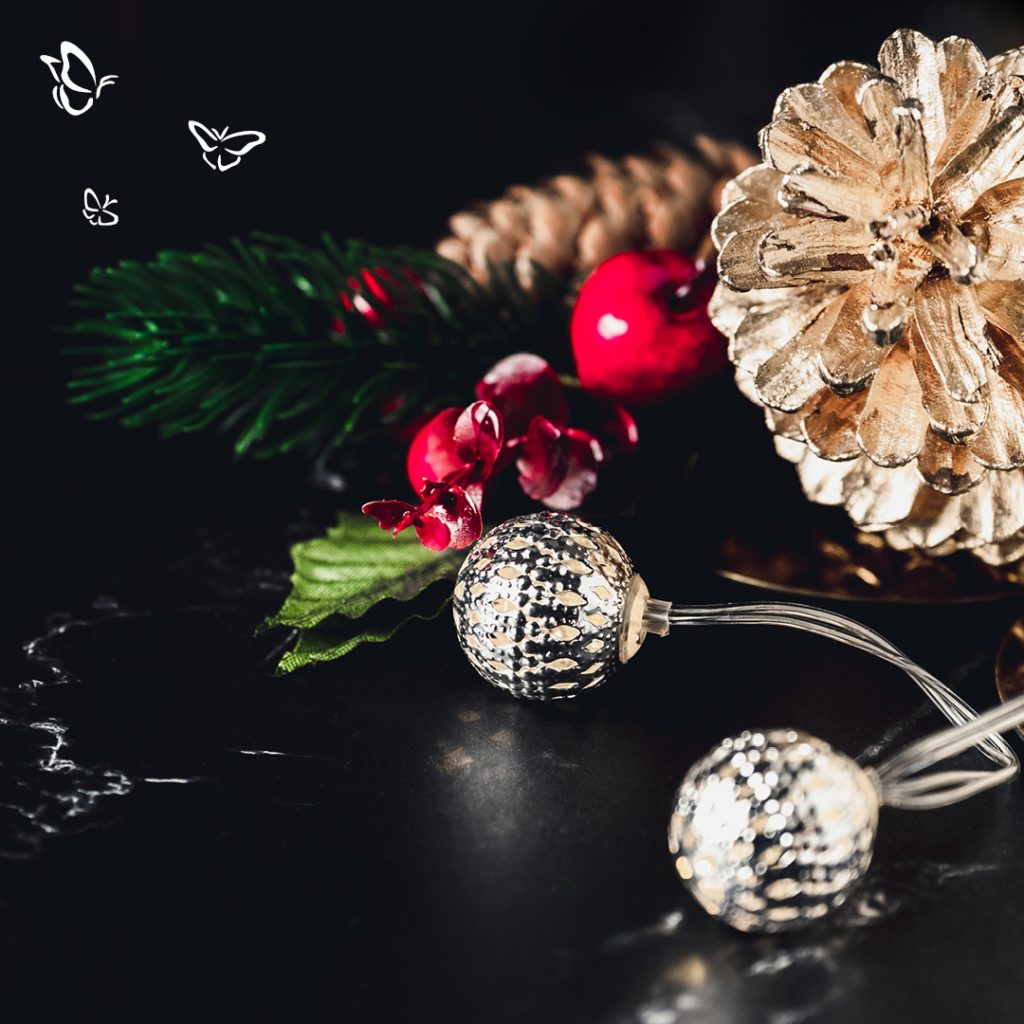 A festive holiday arrangement featuring a gold-painted pinecone and a natural brown pinecone nestled among artificial greenery, red berries, and red flowers. Two spherical string lights with metallic perforations glow warmly in the foreground. The background is a dark, glossy surface that enhances the contrast. Three white butterfly illustrations float in the upper left corner, adding a whimsical touch.