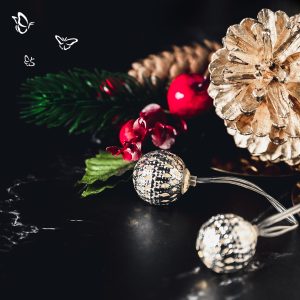 A festive holiday arrangement featuring a gold-painted pinecone and a natural brown pinecone nestled among artificial greenery, red berries, and red flowers. Two spherical string lights with metallic perforations glow warmly in the foreground. The background is a dark, glossy surface that enhances the contrast. Three white butterfly illustrations float in the upper left corner, adding a whimsical touch.