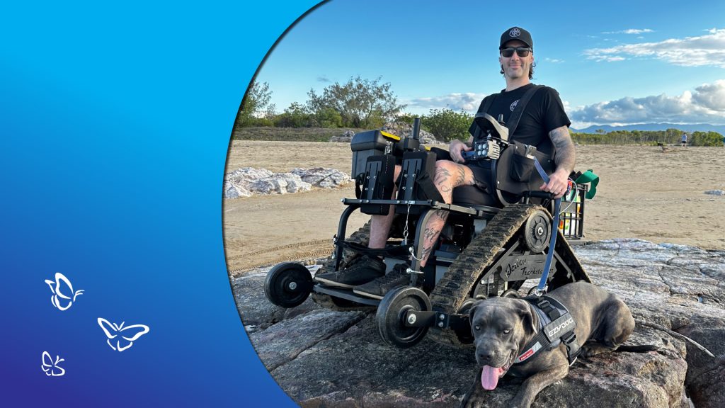 A person sits in an all‑terrain tracked wheelchair on rocky ground in the Australian bush, wearing a black cap, sunglasses, and a black shirt, with tattoos visible on their legs. A dog in an “EZYDOG” harness lies on the rocks in front of them. The scene is surrounded by sandy soil, scattered rocks, and sparse native vegetation under a partly cloudy sky. A blue gradient border with white butterfly illustrations frames the left side of the image, adding a gentle, uplifting accent.