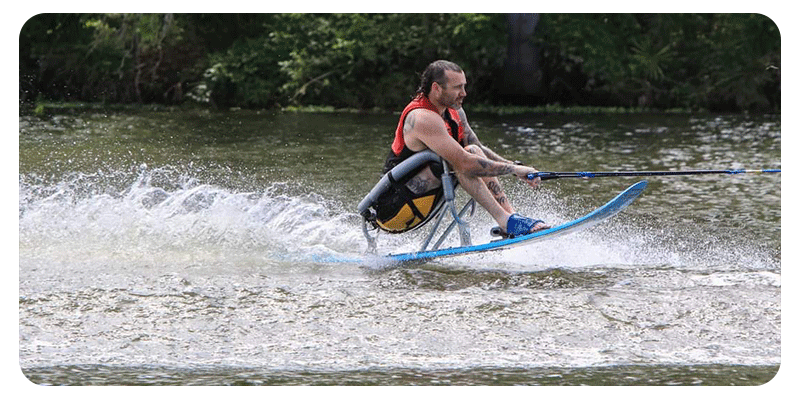 A person is seated in a sit‑ski, wearing a red life vest and gripping a tow rope as they glide across the water. The adaptive ski cuts through the surface, sending up splashes that show speed and motion. The scene highlights adaptive water skiing, with the skier balanced, focused, and fully engaged in the ride.