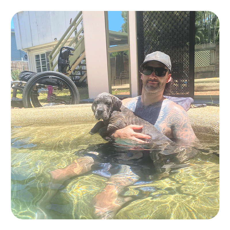 A person sits in a shallow pool holding a small puppy with dark fur and lighter markings. They’re wearing sunglasses and a cap, with tattoos visible on their arms and chest. Sunlight reflects off the water around them. Behind them, a wheelchair, a staircase, and a building with glass doors create an outdoor, relaxed setting.