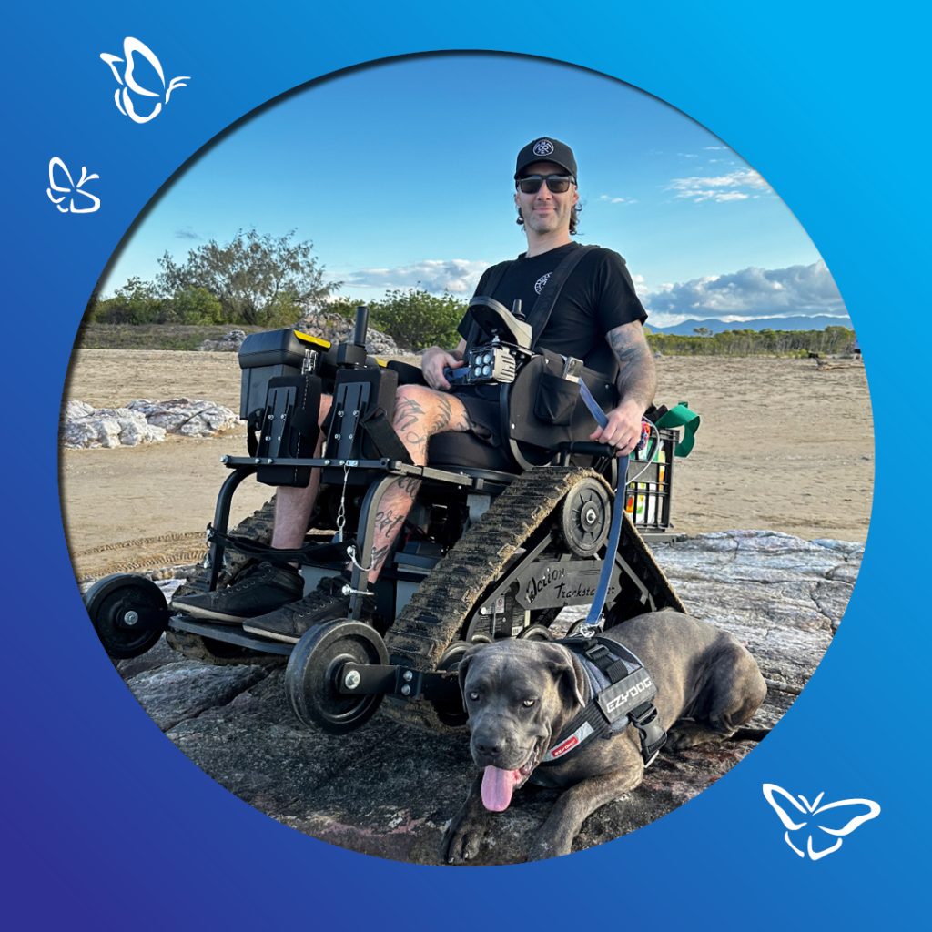 A person sits in an all‑terrain tracked wheelchair on rocky ground in the Australian bush, accompanied by a dog wearing an “EzyDog” harness. The wheelchair’s tank‑like treads rest among stones and dry earth. The person, dressed casually with tattoos, sunglasses, and a cap, appears relaxed and ready to explore. Behind them, native scrub, scattered trees, and an open sky create a rugged bushland backdrop. A blue gradient frame with white butterfly illustrations surrounds the image, adding a sense of lightness and freedom.