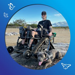 A person sits in an all‑terrain tracked wheelchair on rocky ground in the Australian bush, accompanied by a dog wearing an “EzyDog” harness. The wheelchair’s tank‑like treads rest among stones and dry earth. The person, dressed casually with tattoos, sunglasses, and a cap, appears relaxed and ready to explore. Behind them, native scrub, scattered trees, and an open sky create a rugged bushland backdrop. A blue gradient frame with white butterfly illustrations surrounds the image, adding a sense of lightness and freedom.