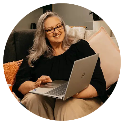 An older person with long gray hair and glasses sits on a couch, smiling as they use a silver Dell laptop. Soft pillows and a knitted orange throw create a cosy, relaxed atmosphere around them.
