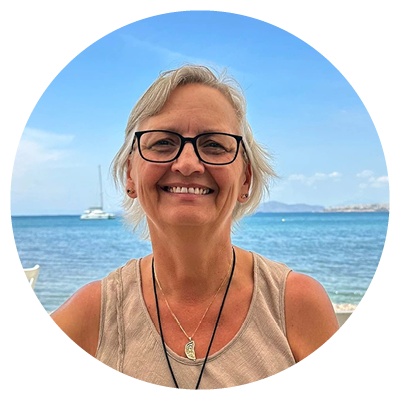 An older person with short gray hair and glasses smiles while standing outdoors in front of a calm blue ocean. They wear a sleeveless beige top and layered necklaces, including one with a leaf-shaped pendant. A sailboat and distant landforms sit under a bright sky with scattered clouds.