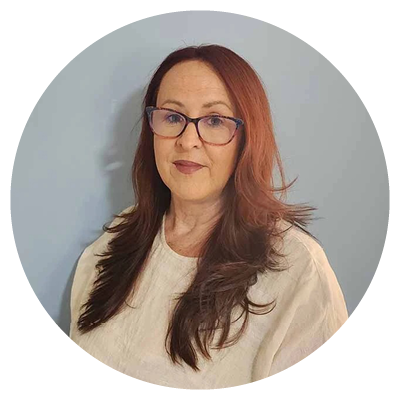 A person with long reddish‑brown hair and glasses sits against a plain light blue background, wearing a light-colored top. The image is cropped in a circular frame, giving it the feel of a simple portrait.
