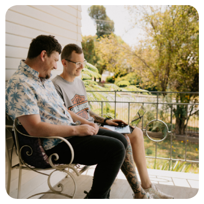 Two people sit on ornate metal chairs on a porch, facing each other in relaxed conversation. One person holds an open laptop on their lap. Behind them, leafy trees and soft greenery create a calm outdoor backdrop. The scene feels casual and friendly, suggesting collaboration or companionship.