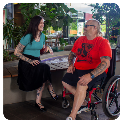 A woman in a teal top and black skirt sits on a bench, smiling as she talks with a man in a wheelchair wearing a red graphic T‑shirt and black shorts. Both have visible tattoos. They sit in a lush, plant‑filled space with a small water feature between them, creating a warm, relaxed atmosphere of connection and accessibility.