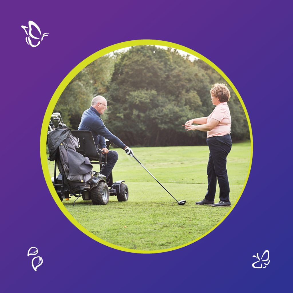 A man using a motorised wheelchair is positioned on a golf course, holding a golf club and preparing to hit a ball. A woman stands in front of him, offering guidance. They’re surrounded by green grass and trees, and the image is framed in a circular border with a purple background decorated with small white butterfly and leaf icons.