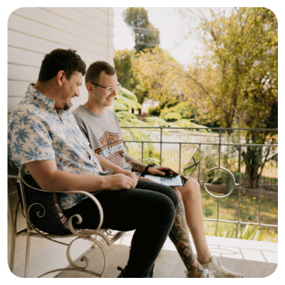 Two people sit on ornate metal chairs on a porch, facing each other in relaxed conversation. One person holds an open laptop on their lap. Behind them, leafy trees and soft greenery create a calm outdoor backdrop. The scene feels casual and friendly, suggesting collaboration or companionship.
