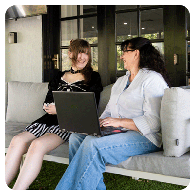 Two people sit together on a light gray outdoor sofa in front of a modern building with large black‑framed windows. One person holds an open laptop on their lap, while the other looks at a smartphone. They appear to be talking as they relax on a grassy patio.