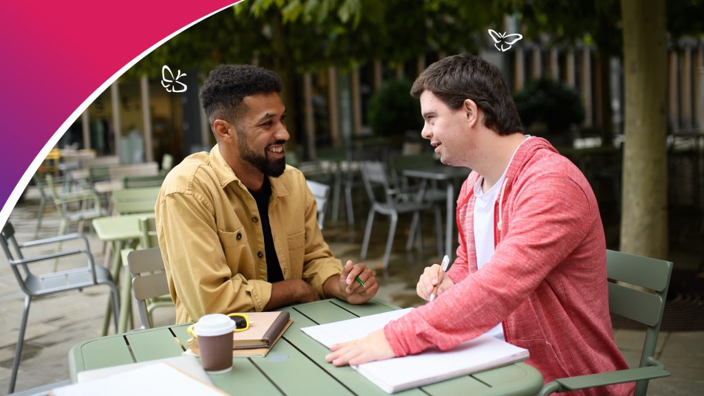 Two people sit together at a green outdoor table, each holding a pen as they talk and work on papers spread out between them. One wears a tan jacket and the other a red hoodie, with notebooks and a coffee cup on the table. They’re in a courtyard with trees and empty tables in the background, suggesting a relaxed, collaborative study or mentoring session.
