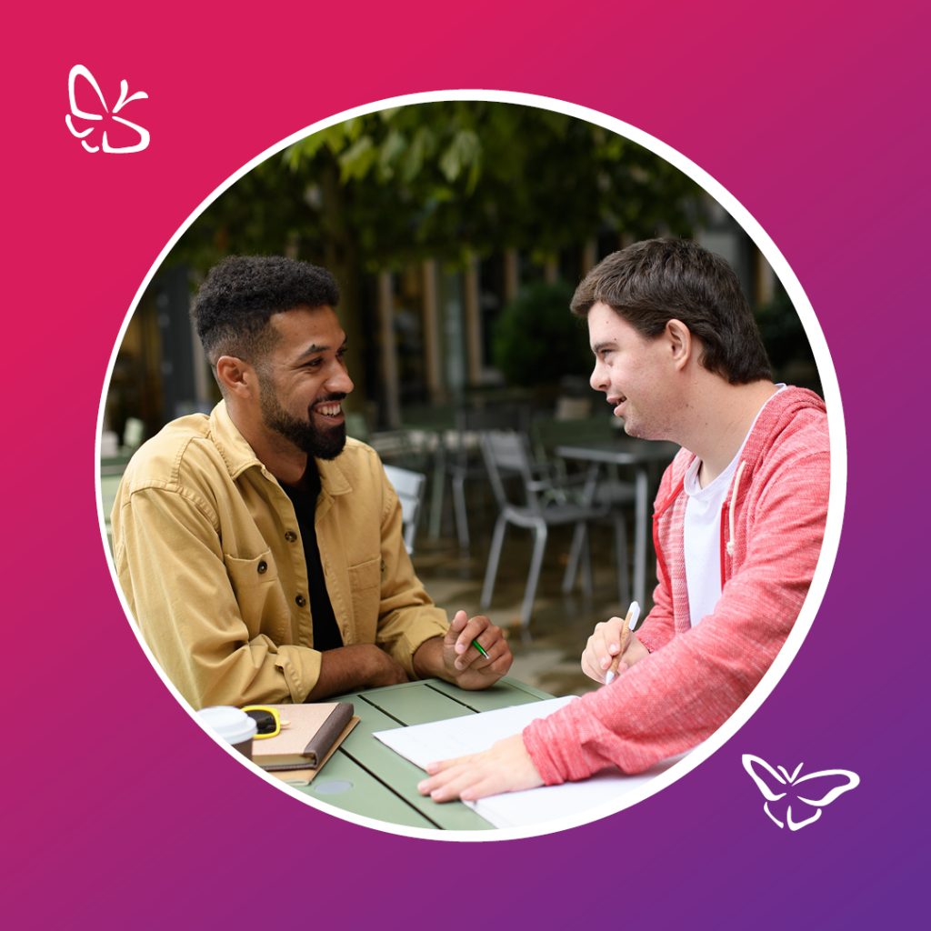 Two people sit at an outdoor table, talking and working together. One person in a tan shirt has a notebook and a cup in front of them, while the other, wearing a red hoodie, holds a pen over a sheet of paper. The setting includes patio seating and greenery, framed within a circular border on a pink‑to‑purple gradient background with small butterfly icons in two corners.
