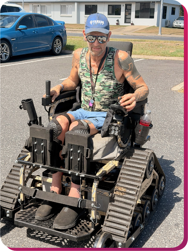 A man wearing a blue Yamaha cap, reflective sunglasses, and a camouflage tank top sits in a rugged, tank‑track wheelchair designed for off‑road mobility. He has tattoos on his arms and a relaxed, confident expression. The wheelchair features heavy‑duty treads, foot supports, and a mounted drink bottle. He’s outdoors on a sunny day, with cars and a white building in the background, symbolising freedom and adaptive innovation.