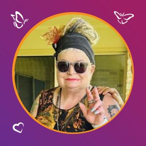 An older person with light hair in a headscarf and sunglasses smiles and gestures with one hand. They wear a patterned sleeveless top and jewelry, framed by a purple background with butterfly and heart icons.