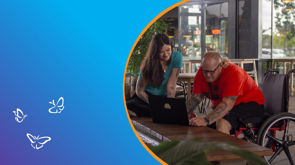 A man using a wheelchair and a woman sit together at a wooden table in a modern café, both smiling as they look at a laptop with a rainbow sticker. The man wears a bright red shirt and black shorts, and the woman wears a teal top. They appear engaged in conversation or collaboration. The scene is framed within a circular orange border on a blue-to-purple gradient background featuring three white butterfly illustrations on the left side, symbolising lightness and inclusion.