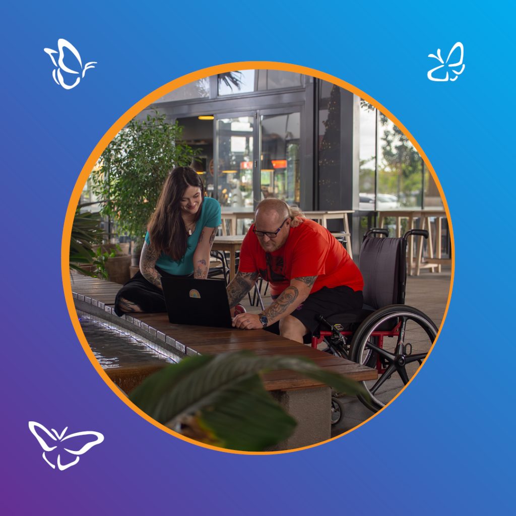 A man using a wheelchair and a woman sit together at a low table beside a shallow water feature, both smiling as they look at a laptop with a rainbow sticker. The man wears a bright red shirt and black shorts, and the woman wears a teal top. The setting appears to be a modern café with plants and large glass windows. The photo is framed within a circular border with an orange outline on a blue-to-purple gradient background, decorated with white butterfly icons.