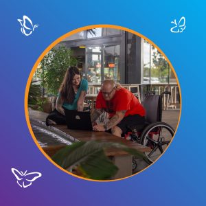 A man using a wheelchair and a woman sit together at a low table beside a shallow water feature, both smiling as they look at a laptop with a rainbow sticker. The man wears a bright red shirt and black shorts, and the woman wears a teal top. The setting appears to be a modern café with plants and large glass windows. The photo is framed within a circular border with an orange outline on a blue-to-purple gradient background, decorated with white butterfly icons.