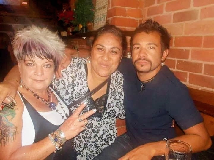 Three people sit close together in a cozy restaurant with brick walls. The person on the left has short spiky hair with light and dark tones, wearing a sleeveless top and jewelry, flashing a peace sign. The person in the middle smiles warmly, wearing a patterned blouse and has an arm around each companion. The person on the right, with short hair and a trimmed beard, wears a dark shirt and necklace, resting one arm on the table beside a glass.